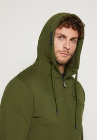 Olivgrüne Zip-Hoodie aus weichem Stoff, mit einer Kapuze mit Kordelzug, gesticktem Logo auf der Brust und kontrastierendem schwarzen Reißverschluss.