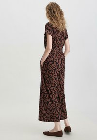 Braun gemustertes Jumpsuit mit kurzen Ärmeln, das eine figurbetonte Taille, Taschen und einen entspannten Schnitt aufweist. Dazu passen braune flache Schuhe aus Wildleder.