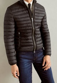 Svart vadderad jacka med horisontell quiltad textur, hög krage och silverfärgad dragkedja. Bärs över en vit skjorta och svart slips, kombinerat med mörka jeans.