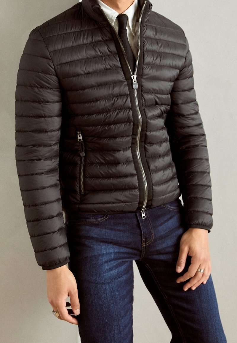 Svart vadderad jacka med horisontell quiltad textur, hög krage och silverfärgad dragkedja. Bärs över en vit skjorta och svart slips, kombinerat med mörka jeans.