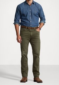 Camisa de mezclilla para hombre en azul oscuro, pantalones verde oliva, cinturón de cuero marrón y botas marrones. La camisa tiene un logo en el bolsillo del pecho.