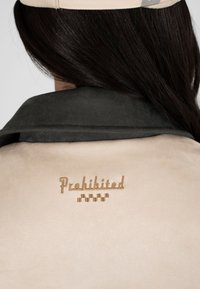 Veste beige avec col sombre portée par une personne aux longs cheveux foncés, brodée du mot "Interdit" et d'un petit motif à carreaux.