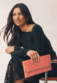 Pochette corail en forme d'enveloppe avec accent métallique, texture lisse. Le modèle porte un haut noir à manches longues et une jupe en dentelle, assise.