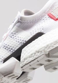 Zapatilla con parte superior de malla blanca, acentos grises y una suela de goma texturizada. Presenta patrones hexagonales distintivos y una lengüeta roja.