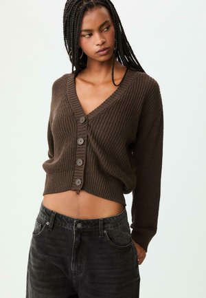 Brown ribbed cardigan with a V-neck, five buttons down the front, and a cropped design. Paired with dark jeans. Textured material.