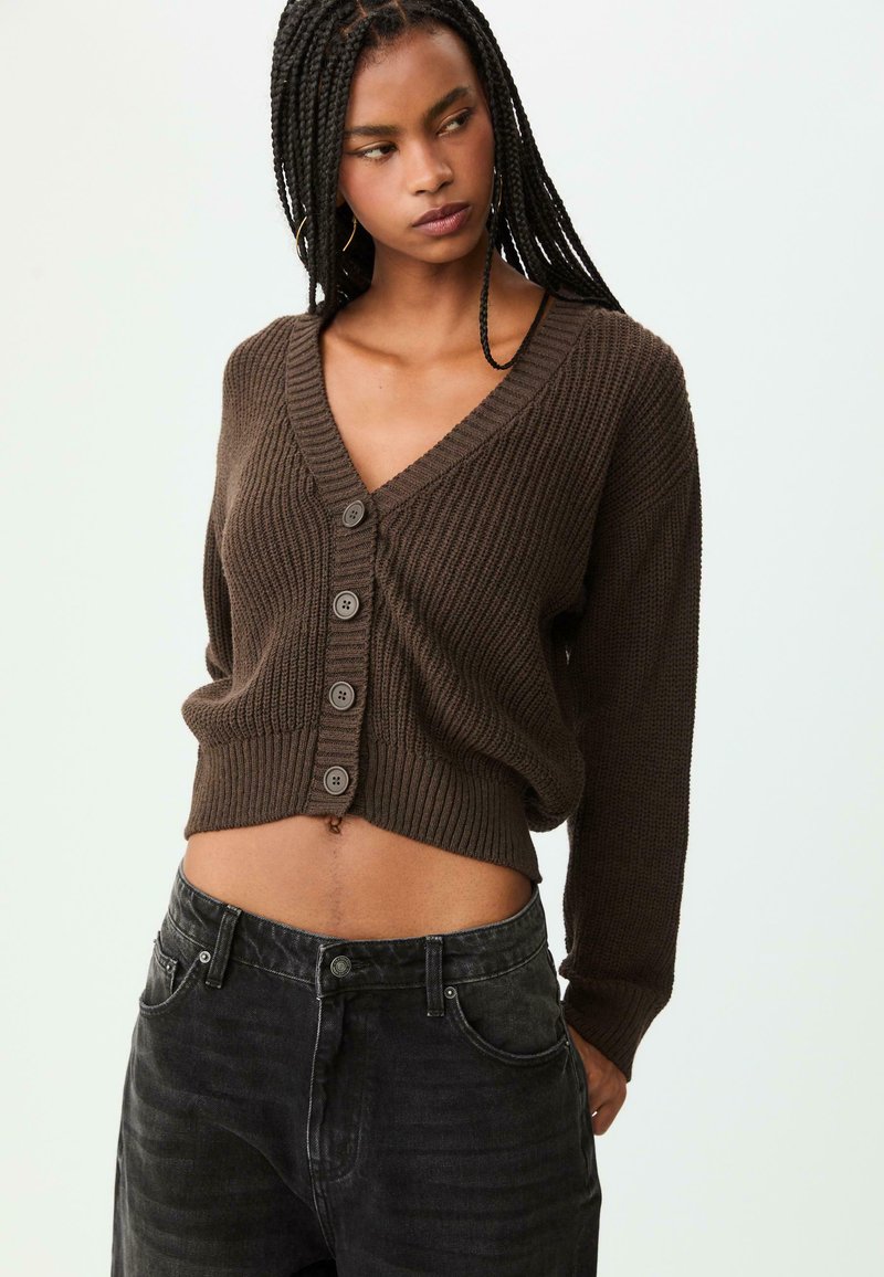 Cardigan marron à côtes avec un col en V, cinq boutons à l'avant et un design court. Associé à un jean foncé. Matériau texturé.