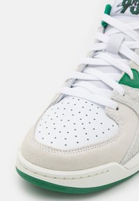 Gros plan sur la pointe d’une basket blanche et beige avec des touches de vert, des lacets blancs et une tige en cuir perforé.