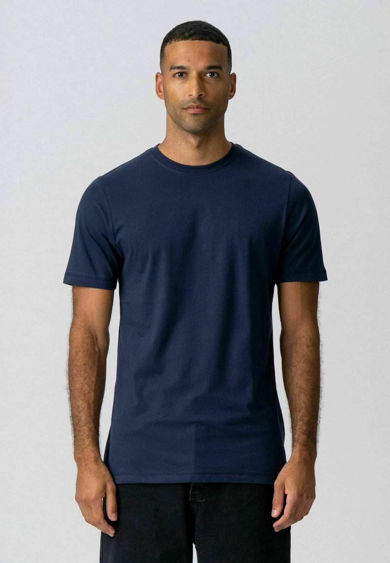 Homme de face portant un t-shirt à manches courtes bleu marine uni et un pantalon noir, sur un fond gris clair.