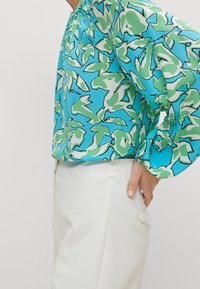 Blouse légère et ample avec un motif coloré de feuilles bleues et vertes, dotée de poignets élastiques et d'un col rond. Associée à un pantalon blanc.