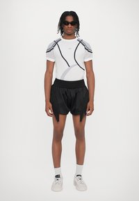 Weißes Kurzarmhemd mit grauen Akzenten und Netzärmeln, kombiniert mit schwarzen Fransen-Shorts. Plateausneaker in gedeckten Farbtönen.