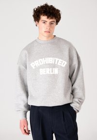 Fiatal férfi, göndör hajjal, szürke pulóvert visel, amelyen a "PROHIBITED BERLIN" felirat található, és sötét redőzött nadrágban áll, egyszínű háttér előtt.