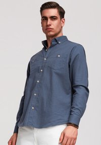 Jeune homme portant une chemise bleue à manches longues boutonnée et un pantalon blanc, debout devant un fond clair uni, portant un bracelet en perles.