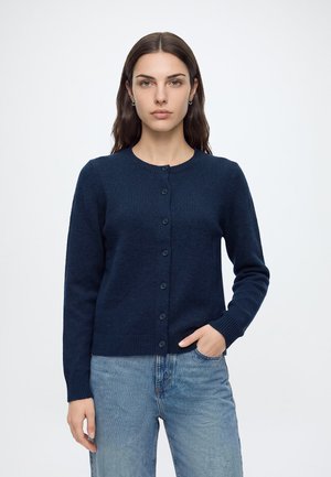 Jeune femme aux cheveux raides et foncés portant un cardigan bleu marine boutonné et un jean bleu clair, debout devant un fond blanc uni.