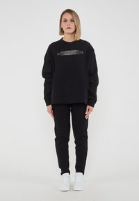 Felpa nera con logo EA7 testurizzato sul petto, abbinata a pantaloni neri e sneakers bianche. Materiale morbido e casual, vestibilità rilassata.