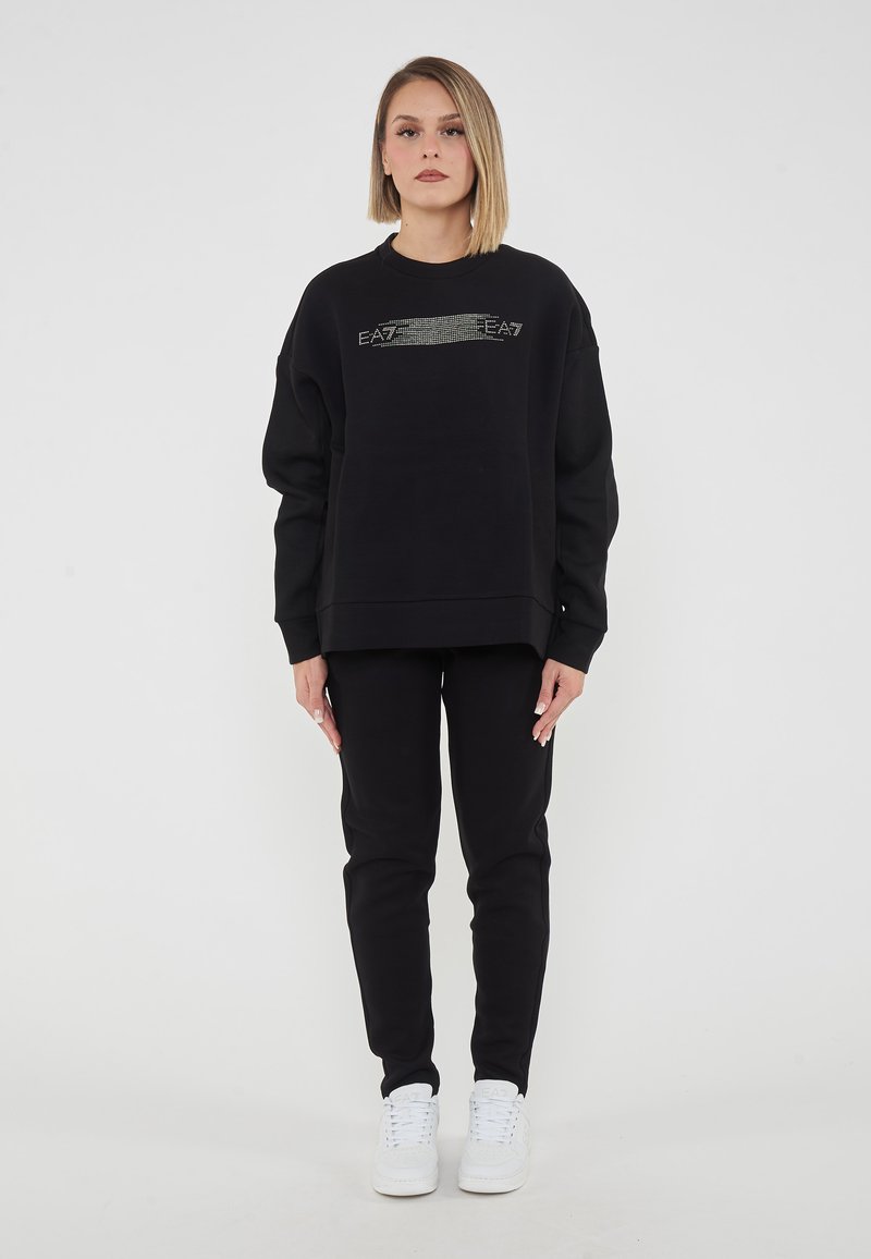 Felpa nera con logo EA7 testurizzato sul petto, abbinata a pantaloni neri e sneakers bianche. Materiale morbido e casual, vestibilità rilassata.