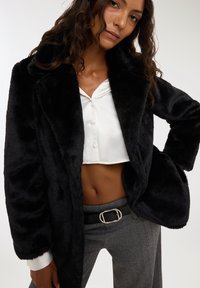 Czarna kurtka z sztucznego futra z szerokim kołnierzem, puszystą teksturą i pełnymi rękawami, noszona na białą, krótką koszulę z guzikami, zestawiona z spodniami w kratę hounds-tooth.