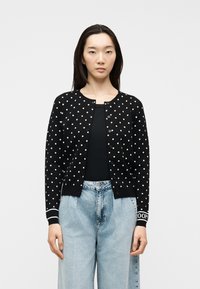 Νεαρή γυναίκα που φοράει μαύρη ζακέτα με κουμπιά και λευκά πουά, μαύρη μπλούζα και ανοιχτό μπλε τζιν με ψηλή μέση, στέκεται μπροστά σε μονόχρωμο φόντο.