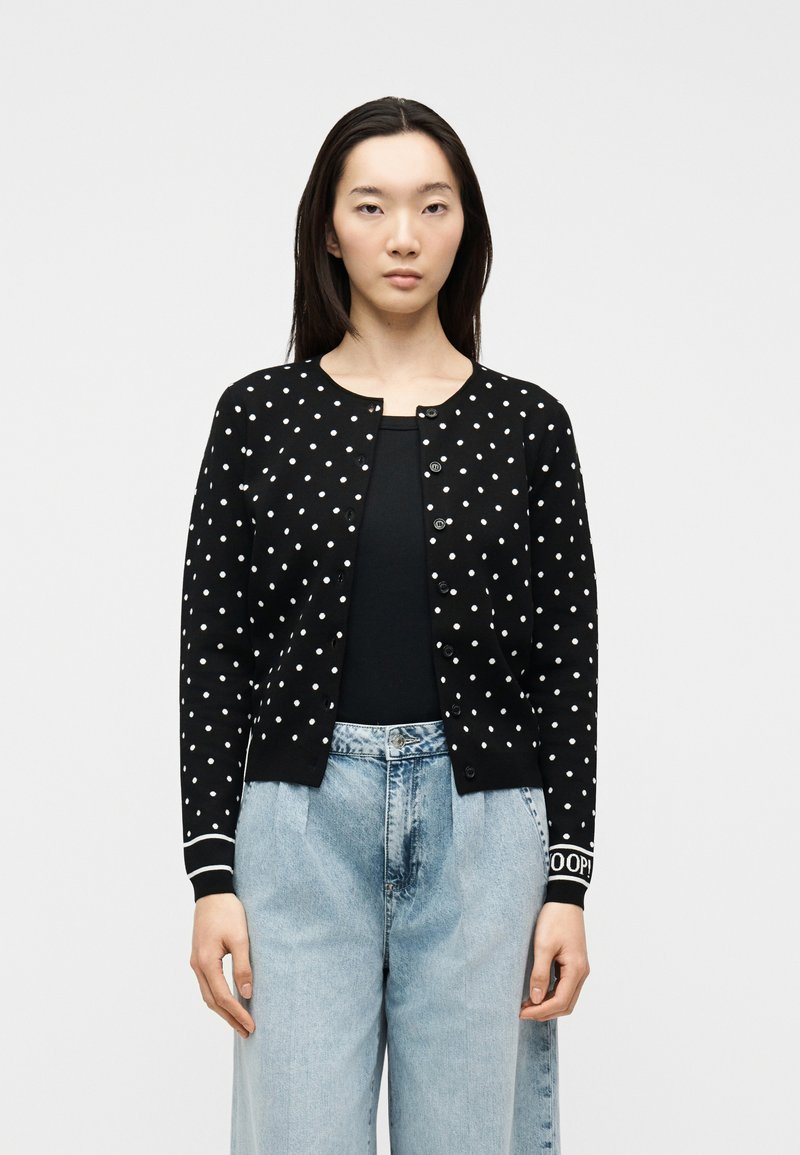 Νεαρή γυναίκα που φοράει μαύρη ζακέτα με κουμπιά και λευκά πουά, μαύρη μπλούζα και ανοιχτό μπλε τζιν με ψηλή μέση, στέκεται μπροστά σε μονόχρωμο φόντο.