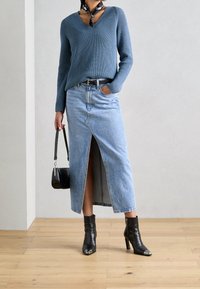 Pull en maille bleu, jupe en denim avec fente frontale, ceinture noire, bottines noires à embouts métalliques, et petit sac à main noir.