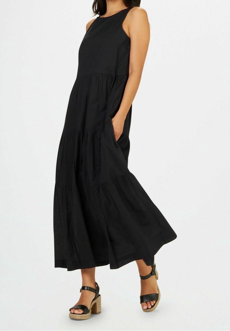 Femme portant une robe midi noire sans manches à volants avec poches et sandales à talons carrés à bride cheville noires sur fond blanc.