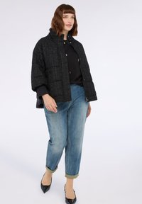 Giacca a vento nera trapuntata con collo alto, caratterizzata da una finitura testurizzata e chiusura frontale con bottoni a pressione, abbinata a jeans azzurri arrotolati.