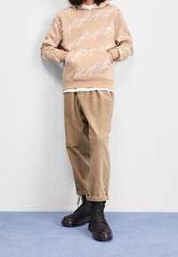 Béžová mikina s bílým potiskem, přes bílou košili. Hnědé kordové kalhoty a černé šněrovací boty dotvářejí outfit.