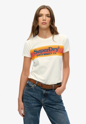 T-shirt crème avec un logo rétro coloré "SuperDry", col rond, manches courtes ajustées, associé à un jean bleu taille haute et une ceinture marron.