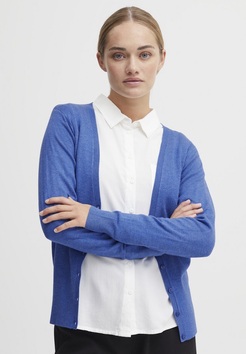 Cardigan bleu avec un col en V, à manches longues, fermeture à boutons et poignets côtelés, porté par-dessus une chemise blanche à col et un pantalon noir.