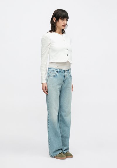 Λευκή κοντή μπλούζα με κουμπιά και μακριά μανίκια, συνδυασμένη με ανοιχτό μπλε άνετο τζιν και ελιάς πράσινα αθλητικά παπούτσια. Απλό φόντο.