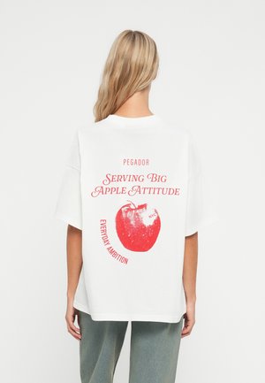 Bijela oversized majica s velikim grafičkim prikazom crvene jabuke na leđima, s natpisom "Serving Big Apple Attitude" u crvenoj i crnoj boji.