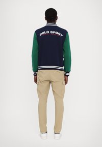 Mörkblå och grön jacka med texten "POLO SPORT" på ryggen, som är kombinerad med beige cargobyxor och vita sneakers.