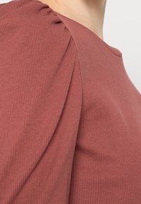 Rostfarbener gerippter Oberteil mit gepufften Ärmeln, runder Ausschnitt und weichem Material, das eine figurbetonte Silhouette betont.