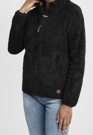 Osoba ubrana w czarną, teksturowaną bluzę polarową rozpinaną na zamek z bocznymi kieszeniami i jasnoniebieskie dżinsy na jednolitym tle.