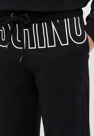 Black sweatpants with elastic waistband, drawstring, and large white lettering. Material appears textured, designed for casual wear.