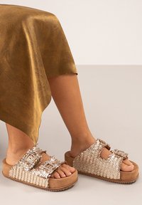 Sandalias metálicas trenzadas con dos hebillas ajustables, suela de plataforma color marrón claro y acabado texturizado. Combinadas con una falda dorada texturizada.