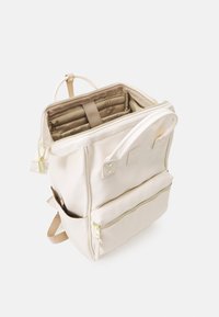 Sac à dos beige avec fermetures éclair dorées, ouverture supérieure montrant des compartiments intérieurs rembourrés, poche avant zippée, poche latérale et doubles poignées de transport.