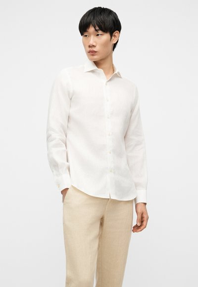 Άνδρας που φοράει λευκό πουκάμισο με μακριά μανίκια και μπεζ παντελόνι, στέκεται με το ένα χέρι στην τσέπη, κοιτώντας προς το πλάι.