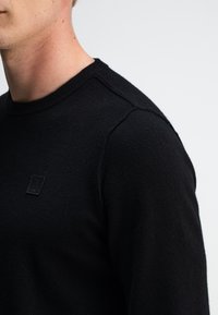 Schwarzer gestrickter Pullover mit Rundhalsausschnitt, der eine strukturierte Oberfläche und ein kleines, geprägtes Logo auf der Brust aufweist. Nahaufnahme von Schulter- und Halsbereich.