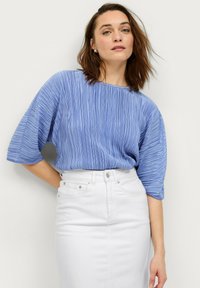 Himmelblaues, plissiertes Blusenshirt mit weiten Ärmeln, kombiniert mit einem weißen, hoch taillierten Rock. Die Bluse weist texturierte Wellenmuster auf.