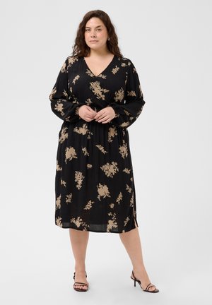 Femme aux cheveux bouclés foncés portant une robe midi noire à imprimé floral beige et manches longues, debout, les mains jointes, portant des sandales noires à talons.