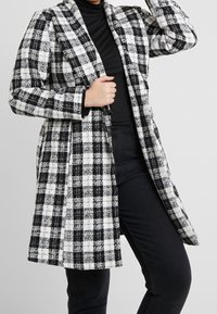 Abrigo de lana en blanco y negro a cuadros, con manga larga, frente abierto y diseño hasta la rodilla. TejidoTexturado con un patrón de cuadros clásico.