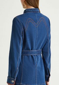 Veste en jean bleu foncé avec col tressé, coutures contrastées et taille ceinturée. Présente un design au dos et un bouton décoratif sur la manche.