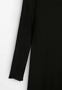 Vêtement à manches longues noir en tissu texturé. Il présente un col rond et un bord légèrement effiloché au niveau du poignet.