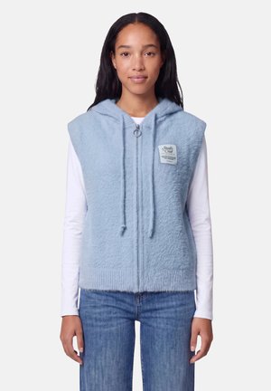 Femme portant un sweat à capuche léger bleu clair sans manches avec une fermeture éclair sur un chemisier blanc à manches longues et un jean bleu, debout devant un fond uni.