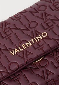 Bordo kožni ručni posao s urezanim uzorkom "VALENTINO" i zlatnim logotipom, s glatkom teksturom i strukturiranim dizajnom.