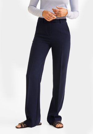 Marineblauwe wijde pantalon van soepel materiaal. Bevat een hoge taille met plooien aan de voorkant. G gedragen met een licht, lang mouwtop en platte sandalen.