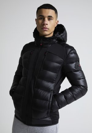 Ung mann iført en svart polstret hettejakke med glidelåslommer, stående med hendene i lommer mot en ensfarget lys bakgrunn.