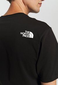 Muž v černém tričku s bílým logem The North Face na horní části zad, mírně otočený na stranu před jednobarevným pozadím.