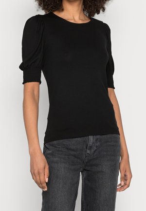 Mujer con una blusa negra ajustada con mangas cortas abullonadas y puños fruncidos combinada con unos vaqueros gris oscuro.