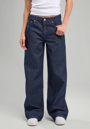 Personne portant un jean large bleu foncé à fines rayures, des baskets blanches et un débardeur court blanc sur un fond gris uni.